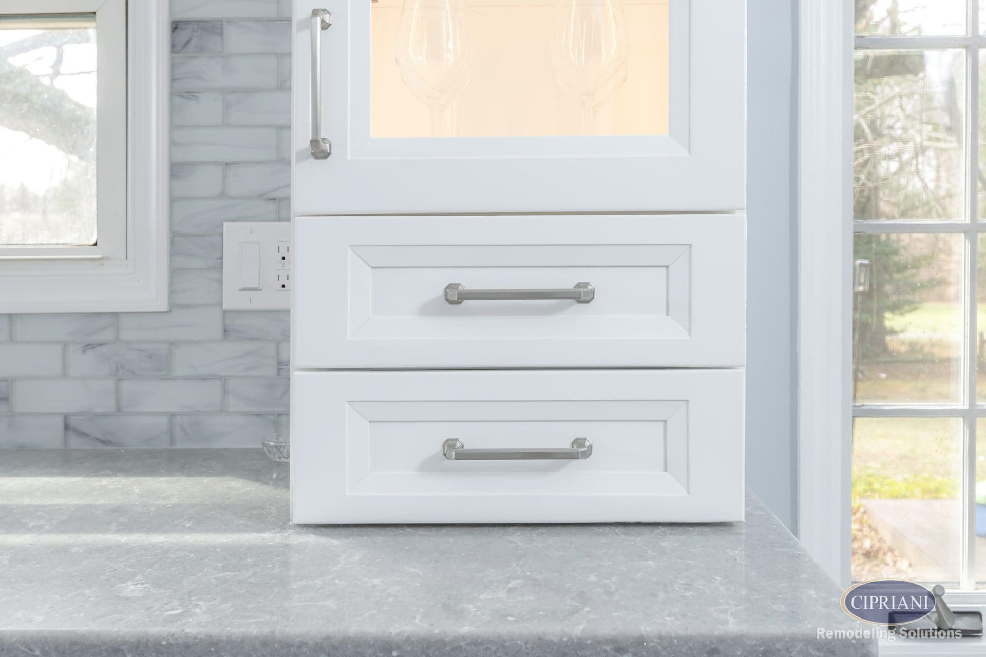 Close-up of white shaker drawers with brushed nickel handles and glass-front cabinet above.