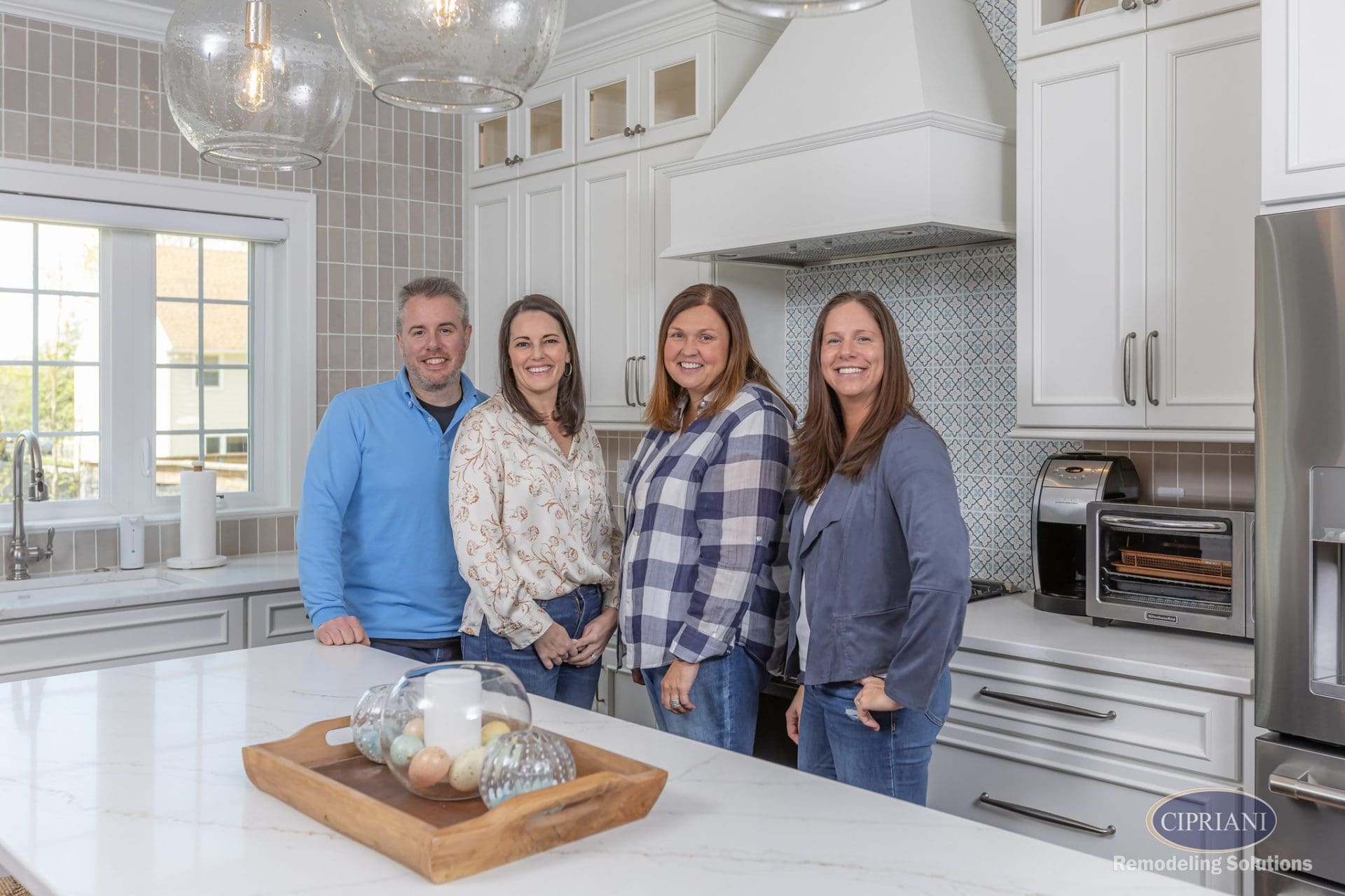 Four people stand smiling in a bright transitional kitchen with white cabinetry, a patterned backsplash, and neutral wall tile.
