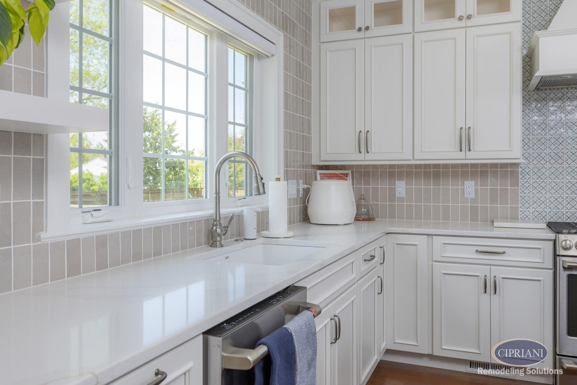 Close-up of white kitchen sink under a large window with surrounding white cabinets and taupe tile backsplash.