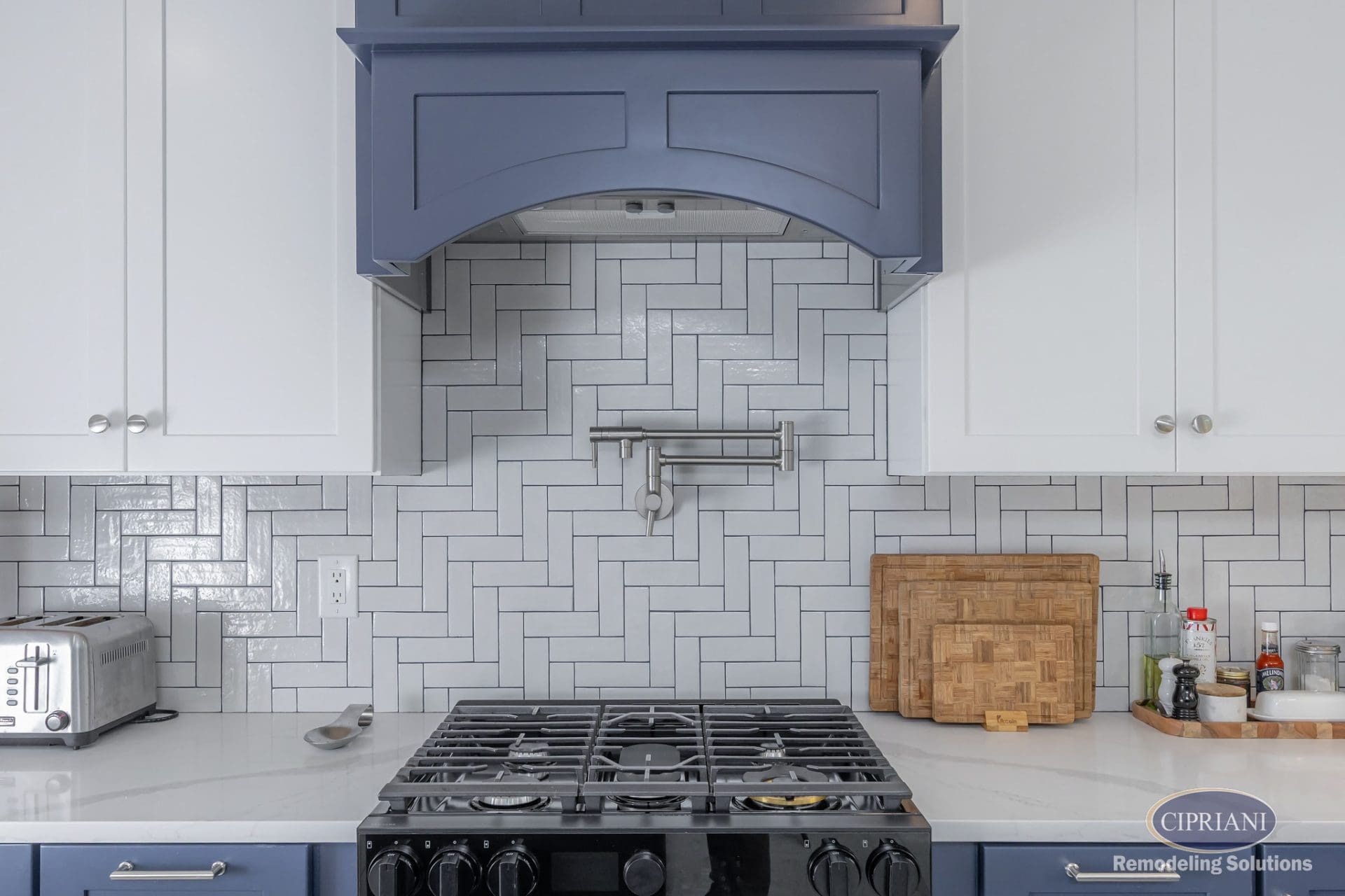 Close-up of kitchen range with pot filler, navy hood, and white herringbone backsplash.