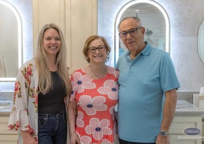 Happy homeowners with designer – A smiling couple stands proudly beside their designer in their newly remodeled bathroom featuring elegant cabinetry and backlit mirrors.