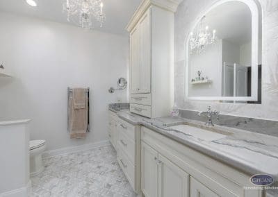 Elegant double vanity – A refined cream-colored vanity with marble countertops and chrome fixtures beneath a glowing arched mirror and crystal chandelier.