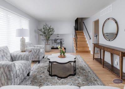 Bright living room with wood stairs – A cozy, sun-filled living room features a patterned area rug, sculptural coffee table, and a wood staircase with updated black railings.