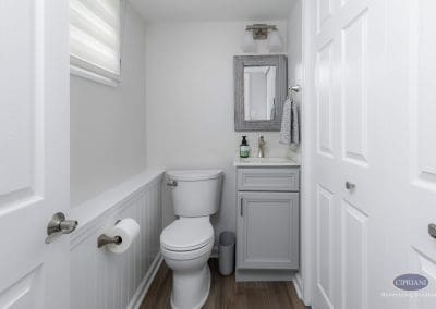 Compact powder room with gray vanity – A small bathroom features a soft gray vanity, brushed nickel fixtures, and a wood-framed mirror above a white countertop.