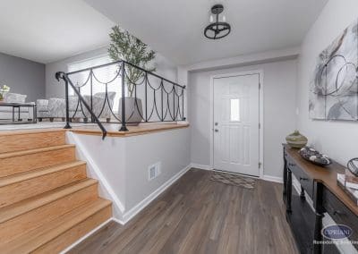 Open Foyer View with Two-Tone Flooring – A bright entryway opens to a cozy upstairs living room, featuring wood steps, iron railing, and layered flooring tones.