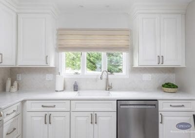 Elegant Sink and Cabinet View – A soft, natural light filters through the window above a sleek undermount sink, surrounded by crisp white cabinetry and a soft herringbone tile backsplash.