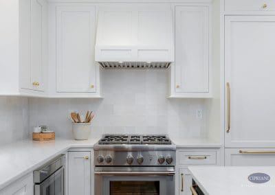 White kitchen cabinets with brass hardware frame a stainless steel range and custom vent hood.