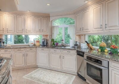 Bright kitchen sink area framed by windows and greenery. Cream cabinetry and granite countertops create a warm, elegant workspace filled with light.
