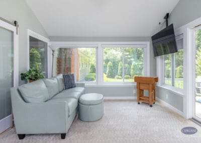 Comfortable sunroom seating with integrated media and backyard views A cozy sunroom seating area featuring a sectional sofa, wall-mounted television, and large windows that frame backyard views while maintaining a relaxed, livable feel.