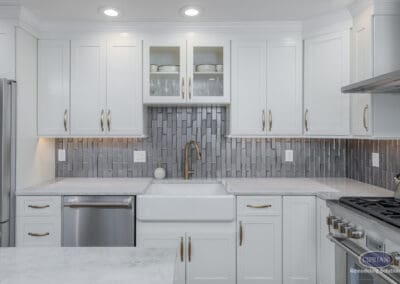 Bright kitchen wall featuring white shaker cabinets, a farmhouse sink, and a textured gray tile backsplash with brass fixtures.