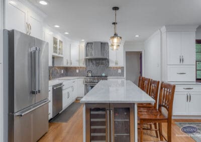 Long kitchen island with marble-look surface, built-in beverage refrigerator, white shaker cabinetry, stainless steel appliances, and brass pendant lighting overhead.