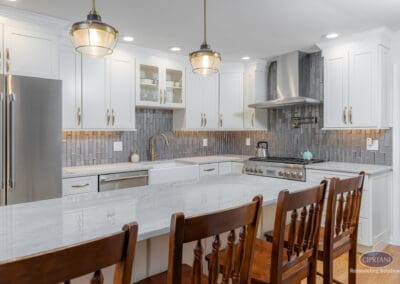 Wide kitchen island with seating, white shaker cabinetry, gray textured tile backsplash, stainless steel range and hood, and brass pendant lighting.