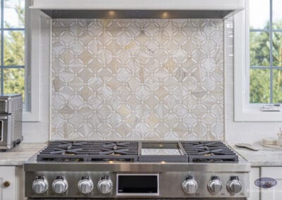 Custom Range and Decorative Tile Backsplash: Professional stainless range centered beneath a sculpted hood with geometric marble tile backsplash framed by windows.