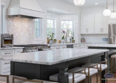Expansive Kitchen Island with Custom Range Wall: Large marble island with seating anchors a bright kitchen featuring white cabinetry, brass hardware, decorative tile backsplash, and custom range hood.