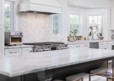 Decorative Tile Range Wall with Custom Hood: Bright kitchen featuring a custom white range hood, patterned tile backsplash, professional gas range, and marble island designed for cooking and entertaining.