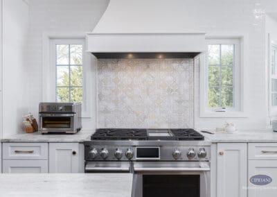 Custom Range Wall with Decorative Tile Detail: Mount Laurel kitchen featuring a professional gas range, custom white range hood, and geometric patterned tile backsplash framed by windows for a bright, timeless focal point.