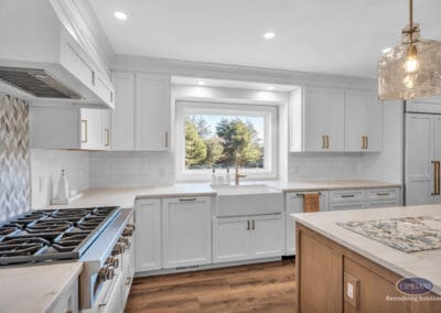 Farmhouse sink under window with white cabinetry, quartz countertops, and brass faucet